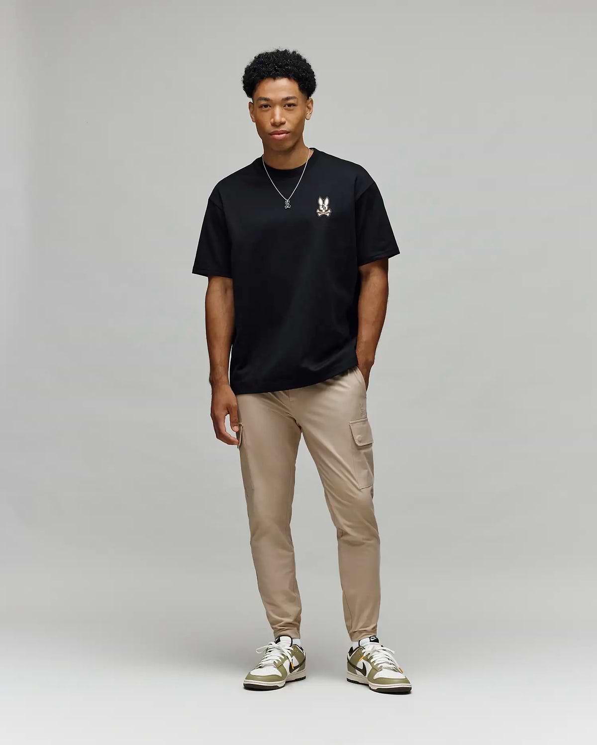 A man stands against a light gray background, wearing Psycho Bunny’s MENS WADE OVERSIZED FIT CHEST TEE (MT0200152) in black with a small chest design, tan cargo pants, white and green sneakers, and a simple necklace.