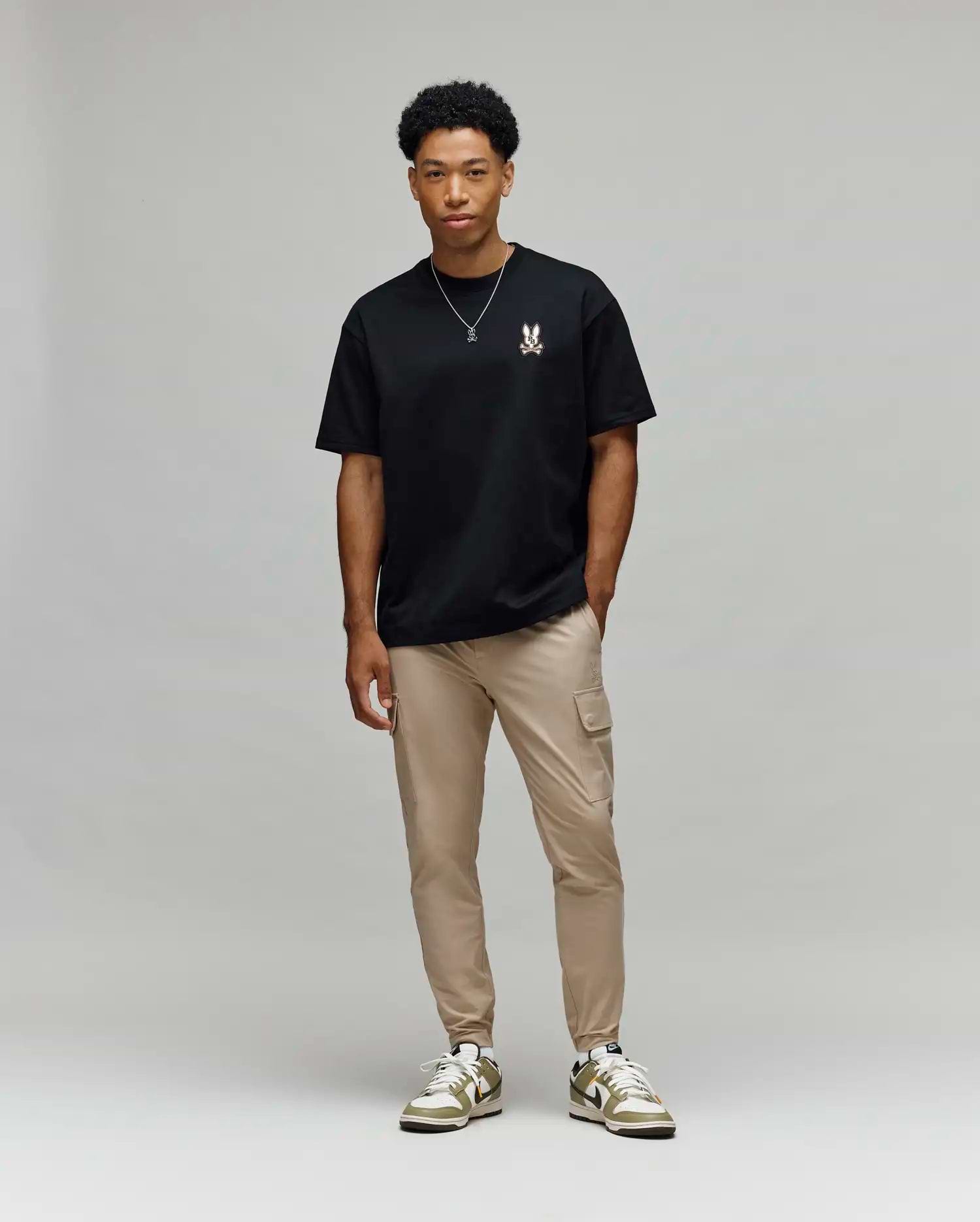 A man stands against a light gray background, wearing Psycho Bunny’s MENS WADE OVERSIZED FIT CHEST TEE (MT0200152) in black with a small chest design, tan cargo pants, white and green sneakers, and a simple necklace.