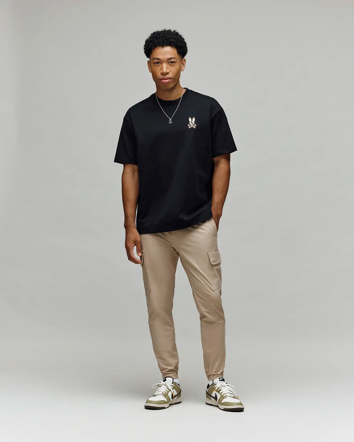 A man stands against a light gray background, wearing Psycho Bunny’s MENS WADE OVERSIZED FIT CHEST TEE (MT0200152) in black with a small chest design, tan cargo pants, white and green sneakers, and a simple necklace.