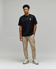 A man stands against a light gray background, wearing Psycho Bunny’s MENS WADE OVERSIZED FIT CHEST TEE (MT0200152) in black with a small chest design, tan cargo pants, white and green sneakers, and a simple necklace.
