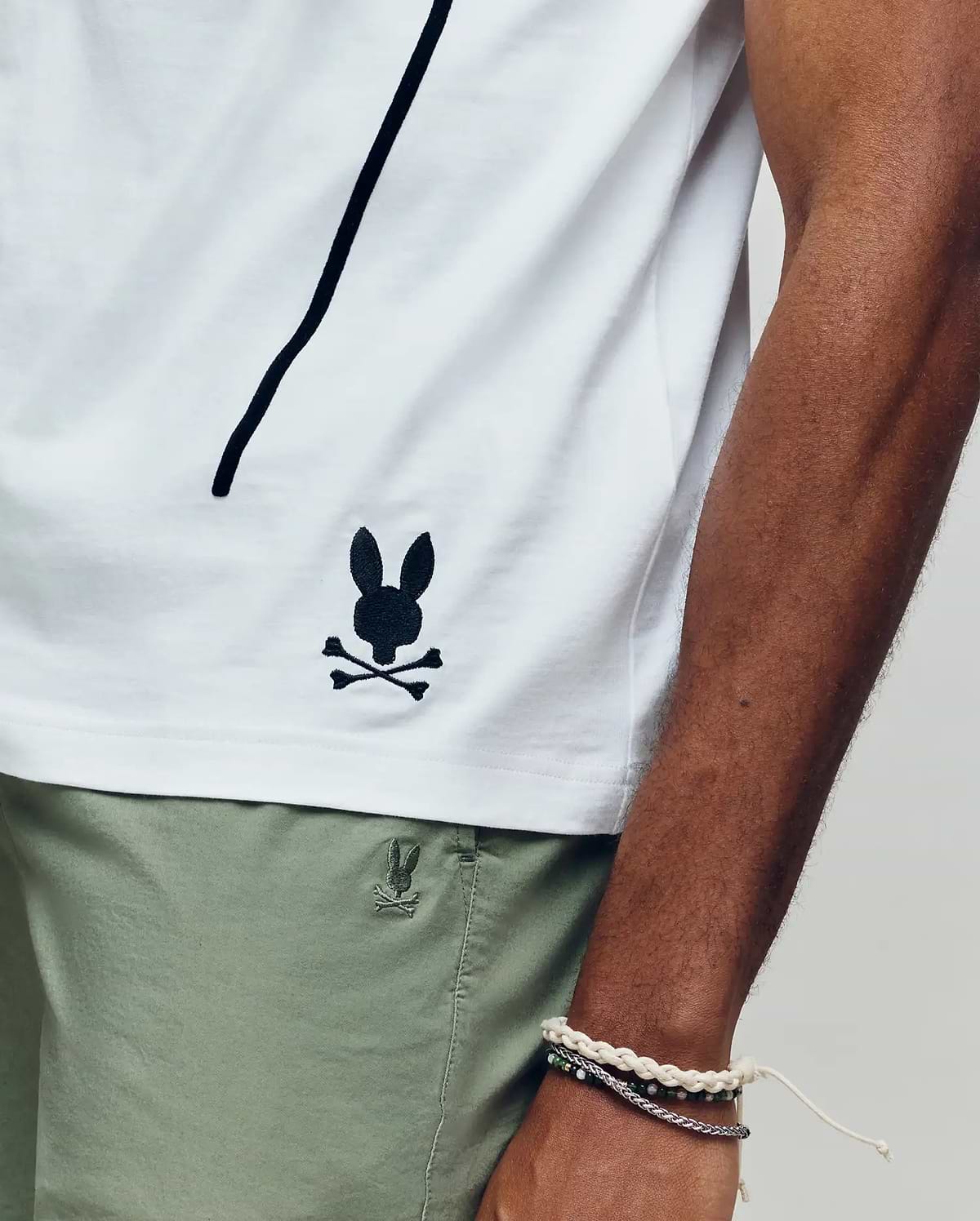 Wearing the Psycho Bunny MENS SOREN GRAPHIC TEE (MT0200180), a 100% cotton white tee with a black bunny and crossbones logo near the hem, and light green shorts, they rest their bracelet-adorned arm along the shirt’s side seam.