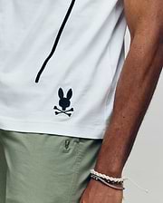 Wearing the Psycho Bunny MENS SOREN GRAPHIC TEE (MT0200180), a 100% cotton white tee with a black bunny and crossbones logo near the hem, and light green shorts, they rest their bracelet-adorned arm along the shirt’s side seam.