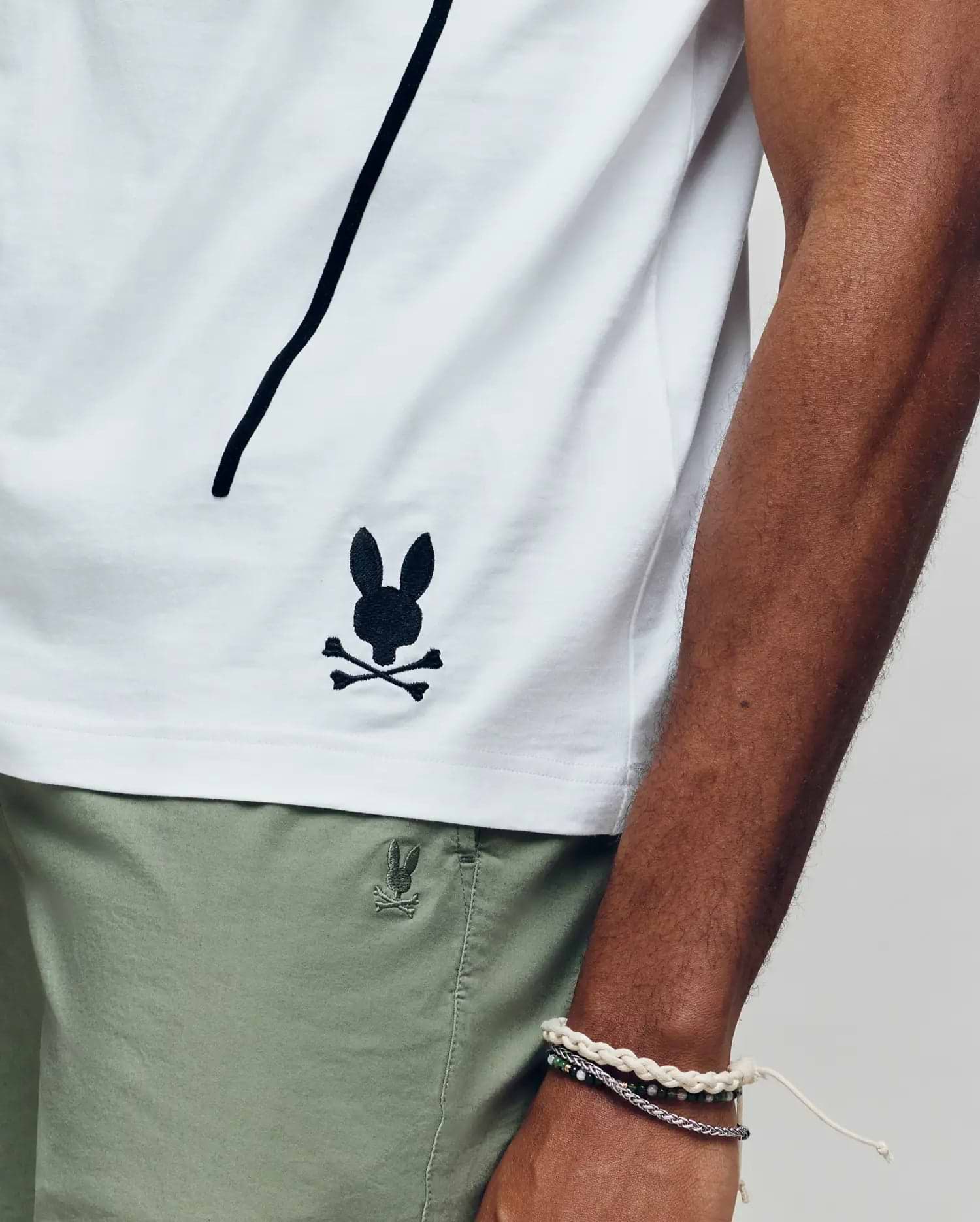 Wearing the Psycho Bunny MENS SOREN GRAPHIC TEE (MT0200180), a 100% cotton white tee with a black bunny and crossbones logo near the hem, and light green shorts, they rest their bracelet-adorned arm along the shirt’s side seam.