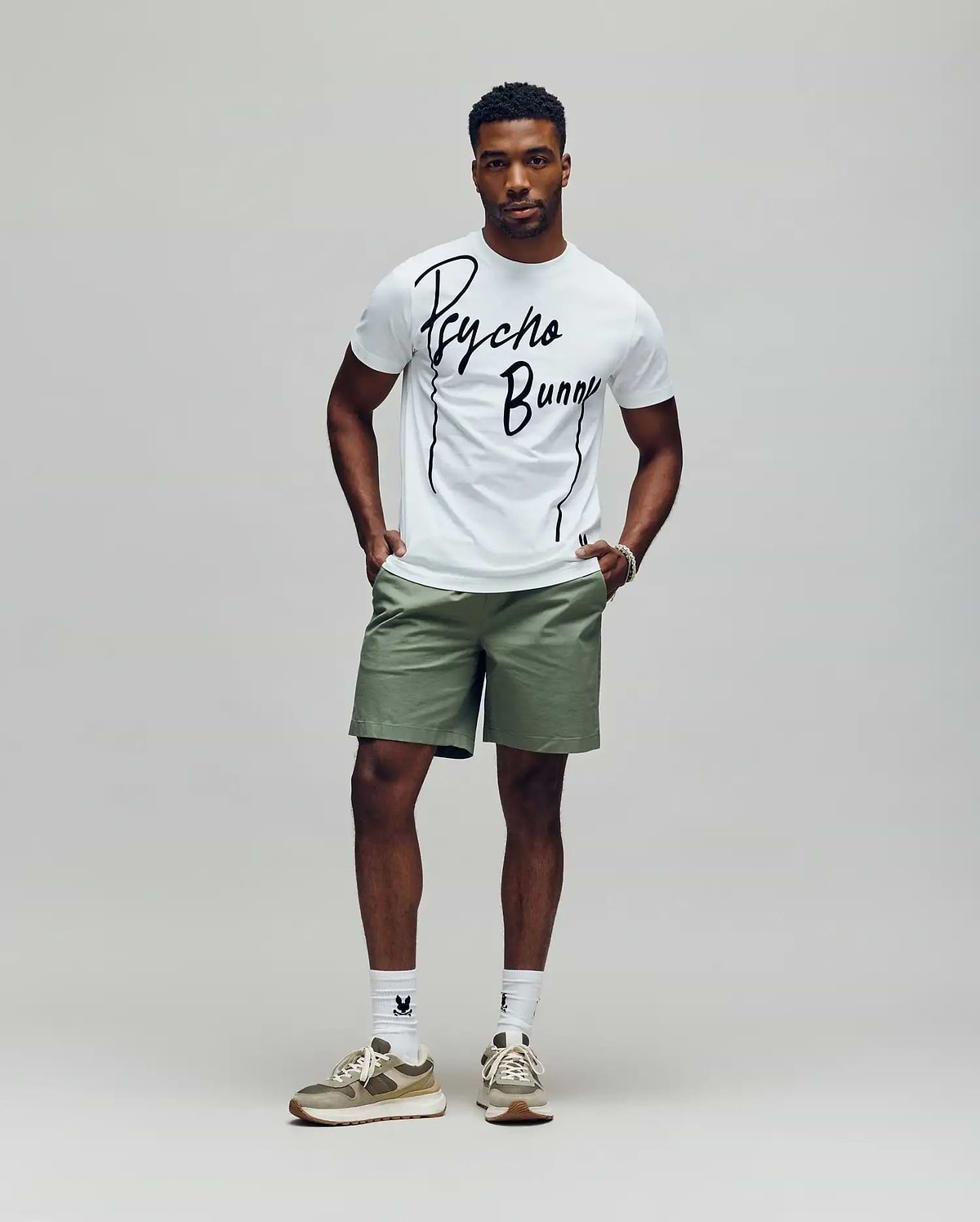 A man stands against a plain background wearing the Psycho Bunny MENS SOREN GRAPHIC TEE (MT0200180) in white, paired with light green shorts, white socks, and beige sneakers. His hands are in his pockets as he looks straight ahead.