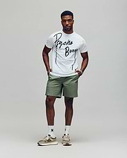 A man stands against a plain background wearing the Psycho Bunny MENS SOREN GRAPHIC TEE (MT0200180) in white, paired with light green shorts, white socks, and beige sneakers. His hands are in his pockets as he looks straight ahead.