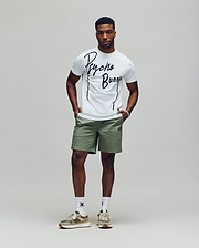 A man stands against a plain background wearing the Psycho Bunny MENS SOREN GRAPHIC TEE (MT0200180) in white, paired with light green shorts, white socks, and beige sneakers. His hands are in his pockets as he looks straight ahead.