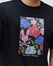 A person wears the MENS ROWEN OVERSIZED TEE (MT0200194) by Psycho Bunny—a relaxed fit black Pima cotton tee with a colorful floral and bunny skull graphic, and "Psycho Bunny" text printed below.