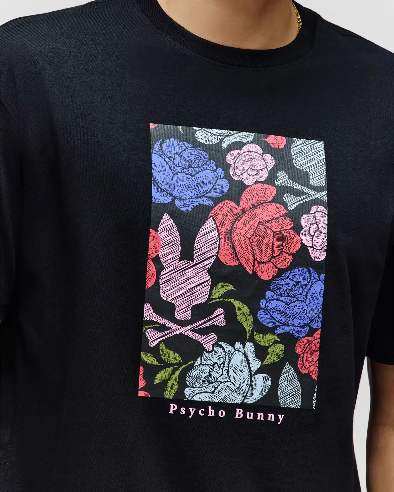 A person wears the MENS ROWEN OVERSIZED TEE (MT0200194) by Psycho Bunny—a relaxed fit black Pima cotton tee with a colorful floral and bunny skull graphic, and "Psycho Bunny" text printed below.