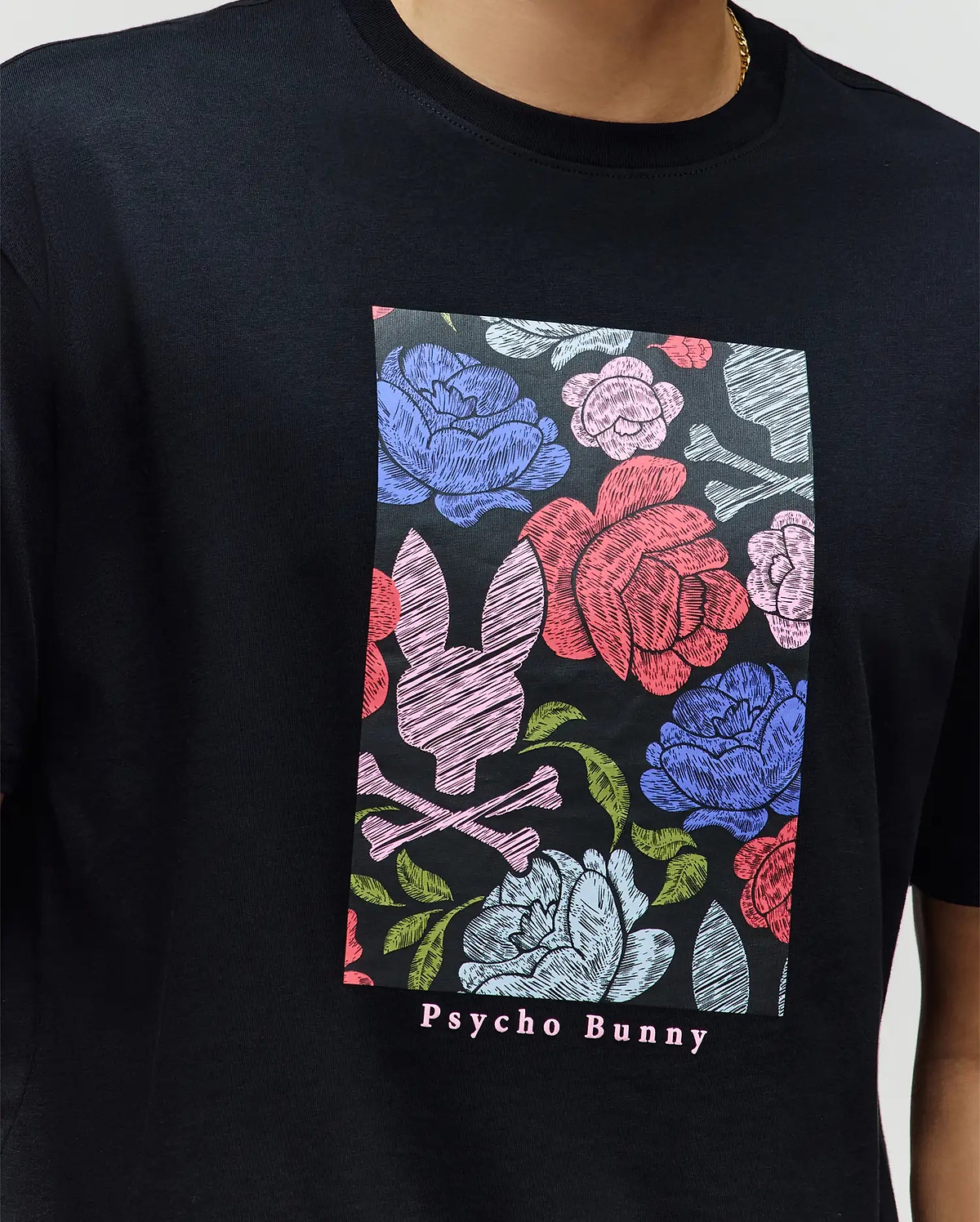A person wears the MENS ROWEN OVERSIZED TEE (MT0200194) by Psycho Bunny—a relaxed fit black Pima cotton tee with a colorful floral and bunny skull graphic, and "Psycho Bunny" text printed below.