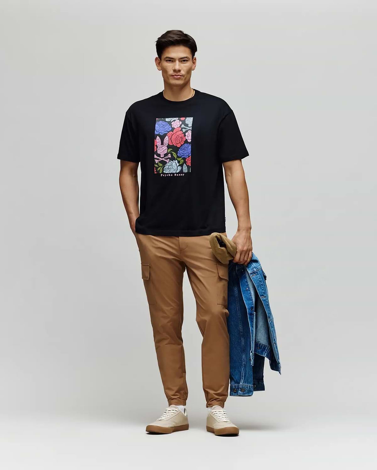 A man smiles at the camera, wearing a black Psycho Bunny MENS ROWEN OVERSIZED TEE (MT0200194) with a floral graphic, tan pants, and beige sneakers, holding a denim jacket and khaki cap in his left hand against a plain backdrop.