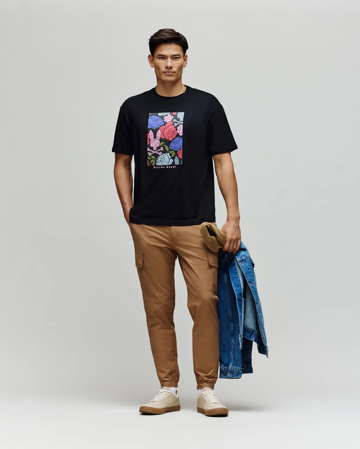 A man smiles at the camera, wearing a black Psycho Bunny MENS ROWEN OVERSIZED TEE (MT0200194) with a floral graphic, tan pants, and beige sneakers, holding a denim jacket and khaki cap in his left hand against a plain backdrop.