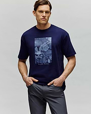 A man stands with hands in pockets against a plain light gray background wearing the Psycho Bunny MENS ROWEN OVERSIZED TEE (MT0200194), a navy blue relaxed-fit shirt featuring a rectangular blue floral design and "Psycho Bunny" text. (1)