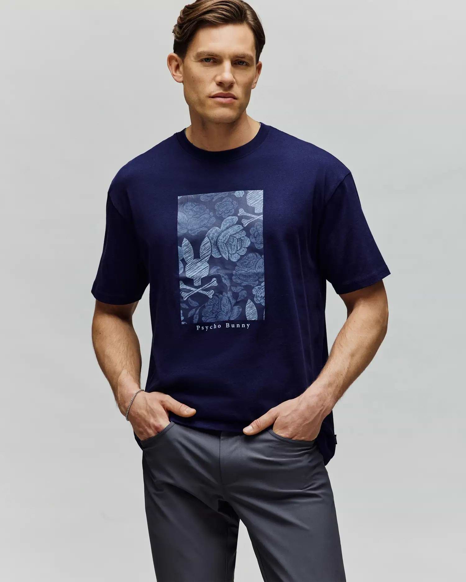 A man stands with hands in pockets against a plain light gray background wearing the Psycho Bunny MENS ROWEN OVERSIZED TEE (MT0200194), a navy blue relaxed-fit shirt featuring a rectangular blue floral design and "Psycho Bunny" text. (1)