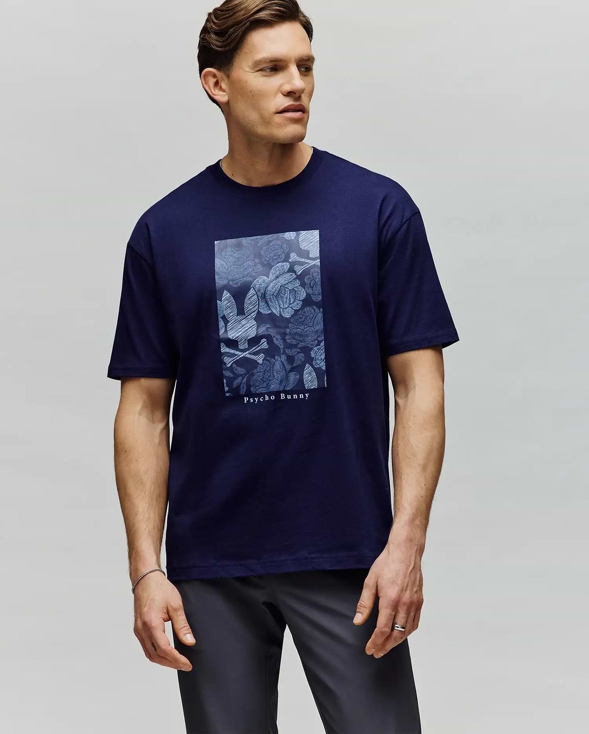 A man with short brown hair stands against a light gray background, wearing the Psycho Bunny MENS ROWEN OVERSIZED TEE (MT0200194)—a navy blue Pima cotton tee with a light blue floral graphic and "Psychic Beauty" text. He looks slightly to the side. (2)