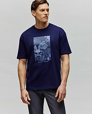 A man with short brown hair stands against a light gray background, wearing the Psycho Bunny MENS ROWEN OVERSIZED TEE (MT0200194)—a navy blue Pima cotton tee with a light blue floral graphic and "Psychic Beauty" text. He looks slightly to the side. (2)