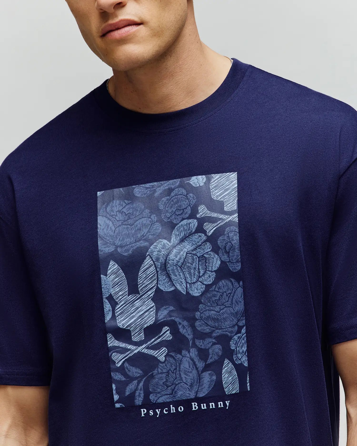 A man wears the Psycho Bunny MENS ROWEN OVERSIZED TEE (MT0200194), a navy blue relaxed fit shirt with a rectangular floral graphic and stylized bunny skull and crossbones on the chest. (3)