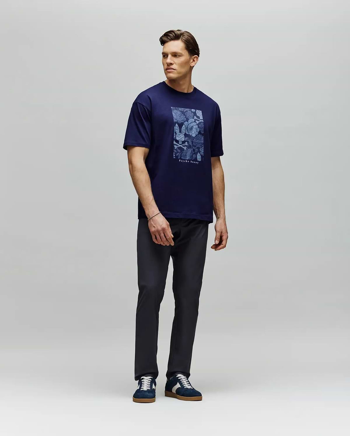 A man stands against a light gray backdrop wearing the Psycho Bunny MENS ROWEN OVERSIZED TEE (MT0200194) in navy blue, paired with dark pants and navy sneakers. He looks to his left with his arms relaxed at his sides. (4)