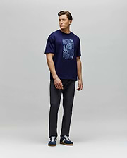 A man stands against a light gray backdrop wearing the Psycho Bunny MENS ROWEN OVERSIZED TEE (MT0200194) in navy blue, paired with dark pants and navy sneakers. He looks to his left with his arms relaxed at his sides. (4)