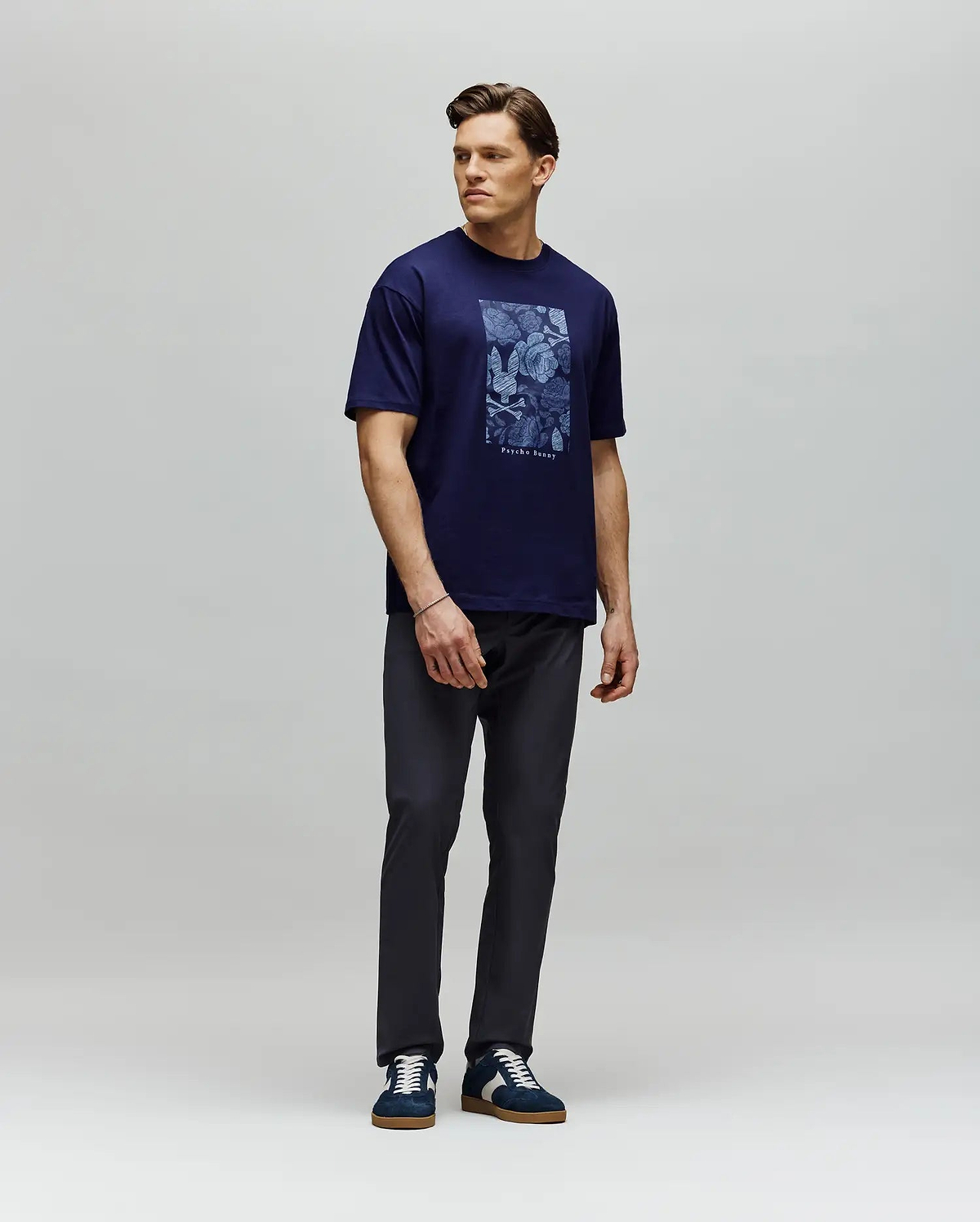 A man stands against a light gray backdrop wearing the Psycho Bunny MENS ROWEN OVERSIZED TEE (MT0200194) in navy blue, paired with dark pants and navy sneakers. He looks to his left with his arms relaxed at his sides. (4)