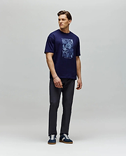 A man stands against a light gray backdrop wearing the Psycho Bunny MENS ROWEN OVERSIZED TEE (MT0200194) in navy blue, paired with dark pants and navy sneakers. He looks to his left with his arms relaxed at his sides. (4)