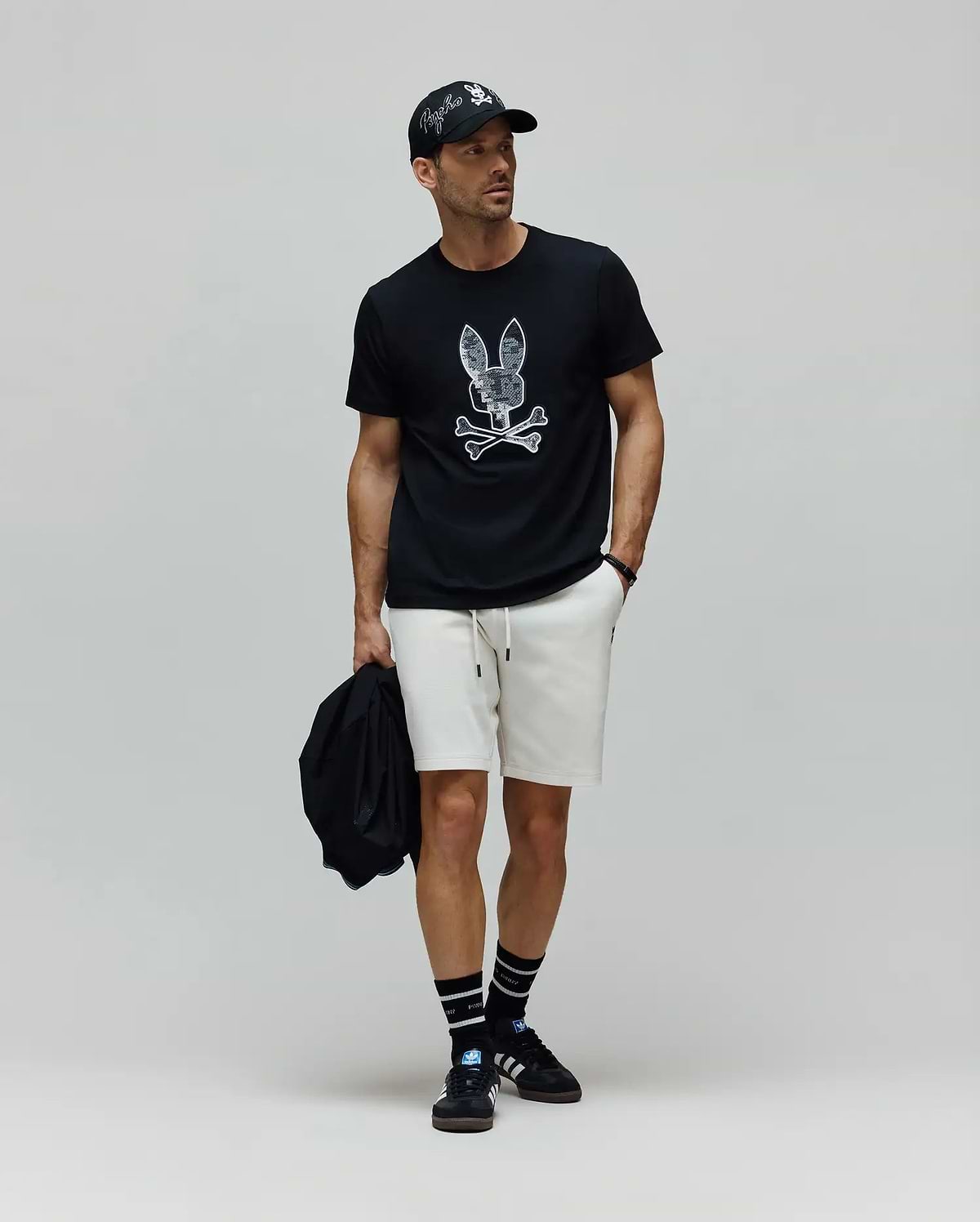 Mens Olivier Graphic Tee in black jersey with a large white distressed Psycho Bunny skull graphic, paired with cream relaxed-fit knit shorts, a black cap featuring an embroidered white Psycho Bunny logo, and black striped crew socks. (5)