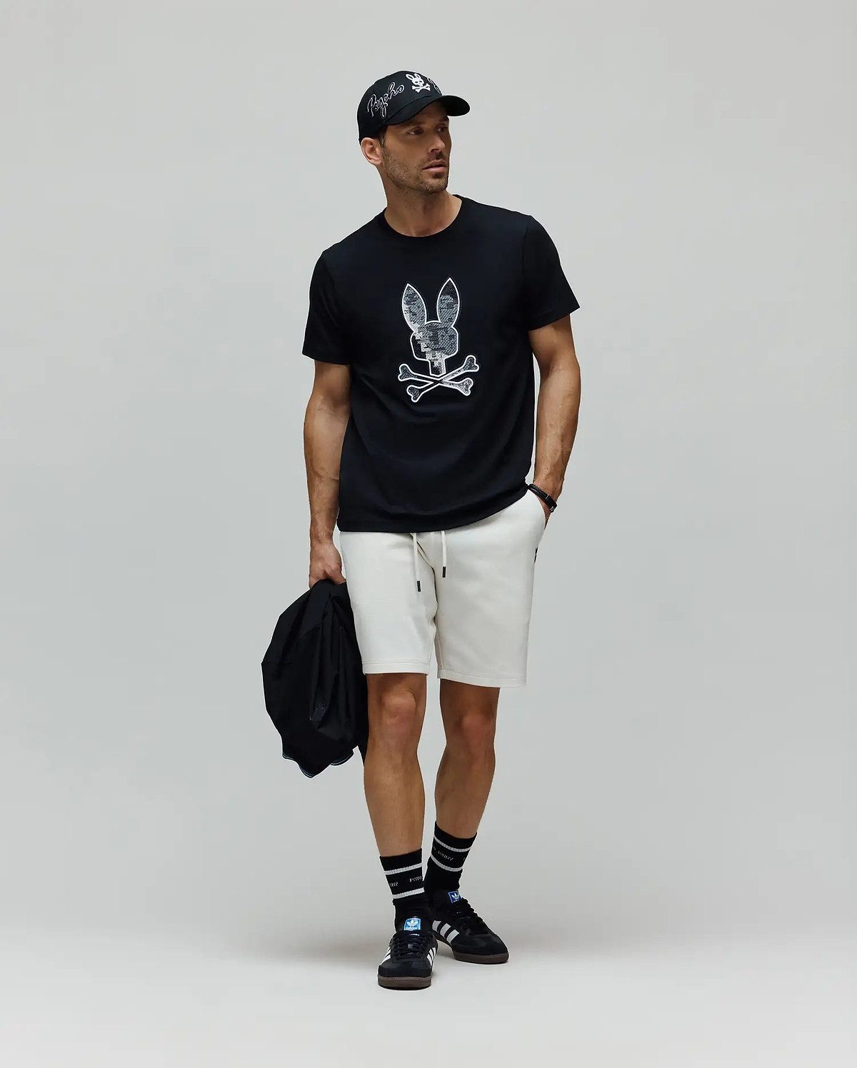Mens Olivier Graphic Tee in black jersey with a large white distressed Psycho Bunny skull graphic, paired with cream relaxed-fit knit shorts, a black cap featuring an embroidered white Psycho Bunny logo, and black striped crew socks. (5)