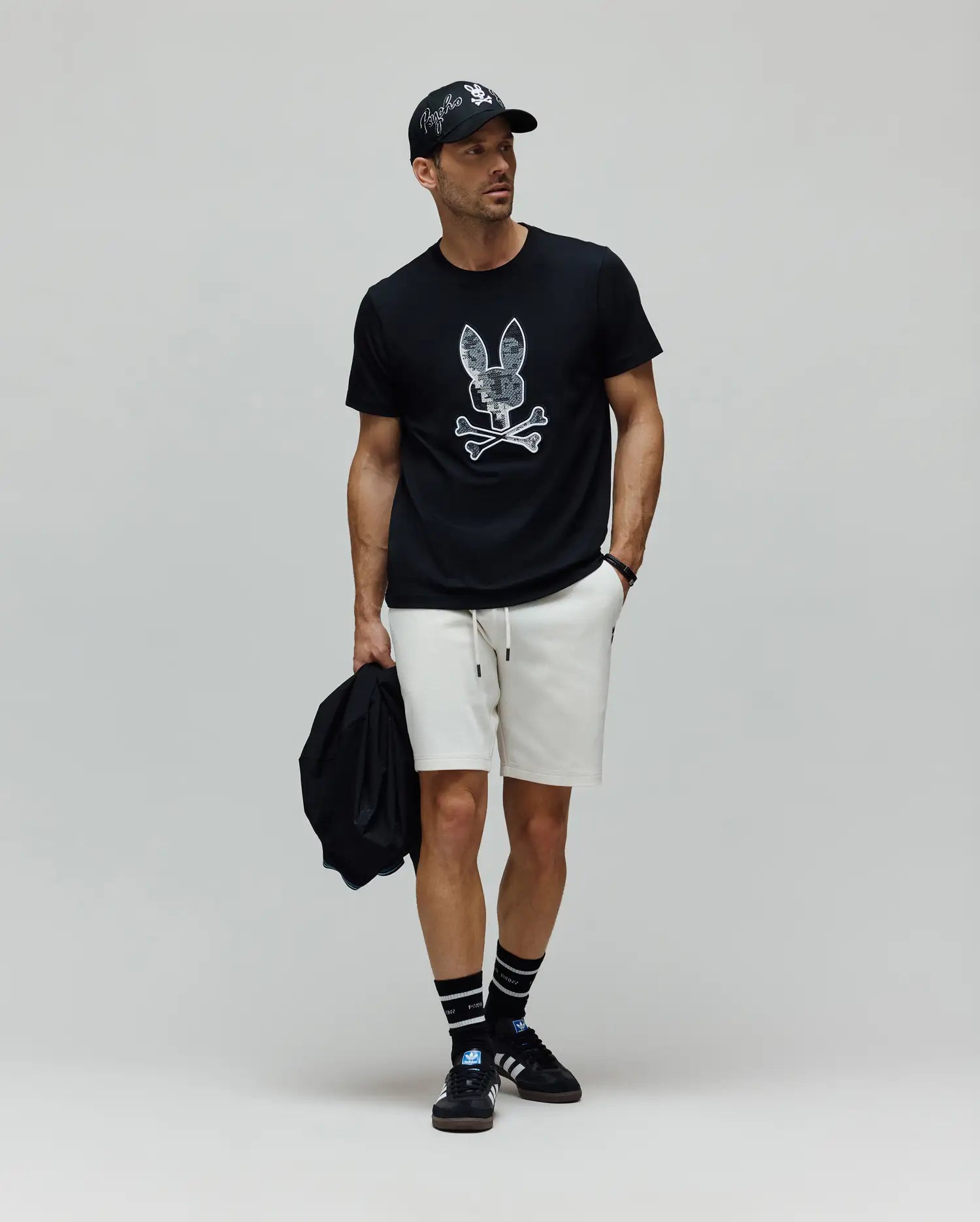 Mens Olivier Graphic Tee in black jersey with a large white distressed Psycho Bunny skull graphic, paired with cream relaxed-fit knit shorts, a black cap featuring an embroidered white Psycho Bunny logo, and black striped crew socks. (5)