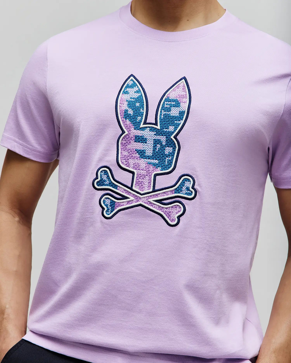 Mens Olivier Graphic Tee, a classic fit lavender jersey knit t-shirt with a large embroidered Psycho Bunny skull logo on the chest, featuring a pixelated blue and purple pattern with a dark blue outline. (3)