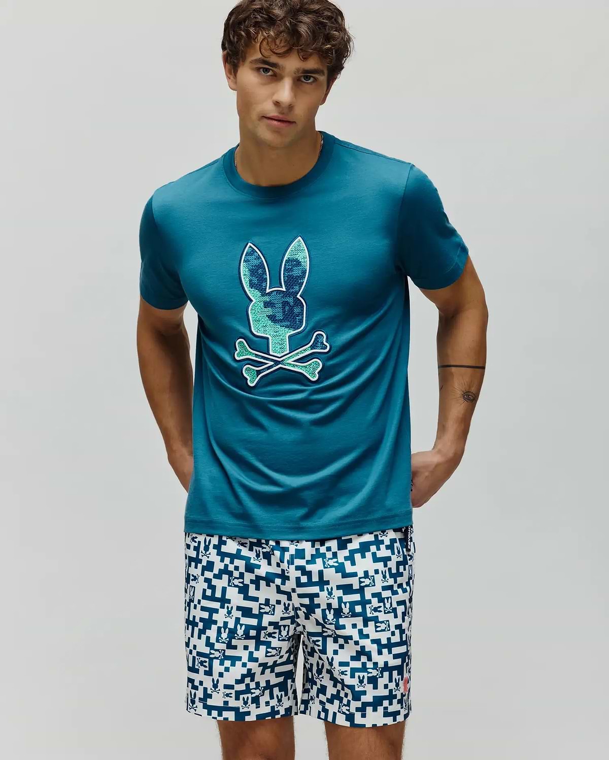 Mens Olivier Graphic Tee, a classic fit teal jersey t-shirt with a large pixelated blue and green Psycho Bunny skull and crossbones chest graphic. White shorts feature an all-over blue geometric bunny pattern. (2)