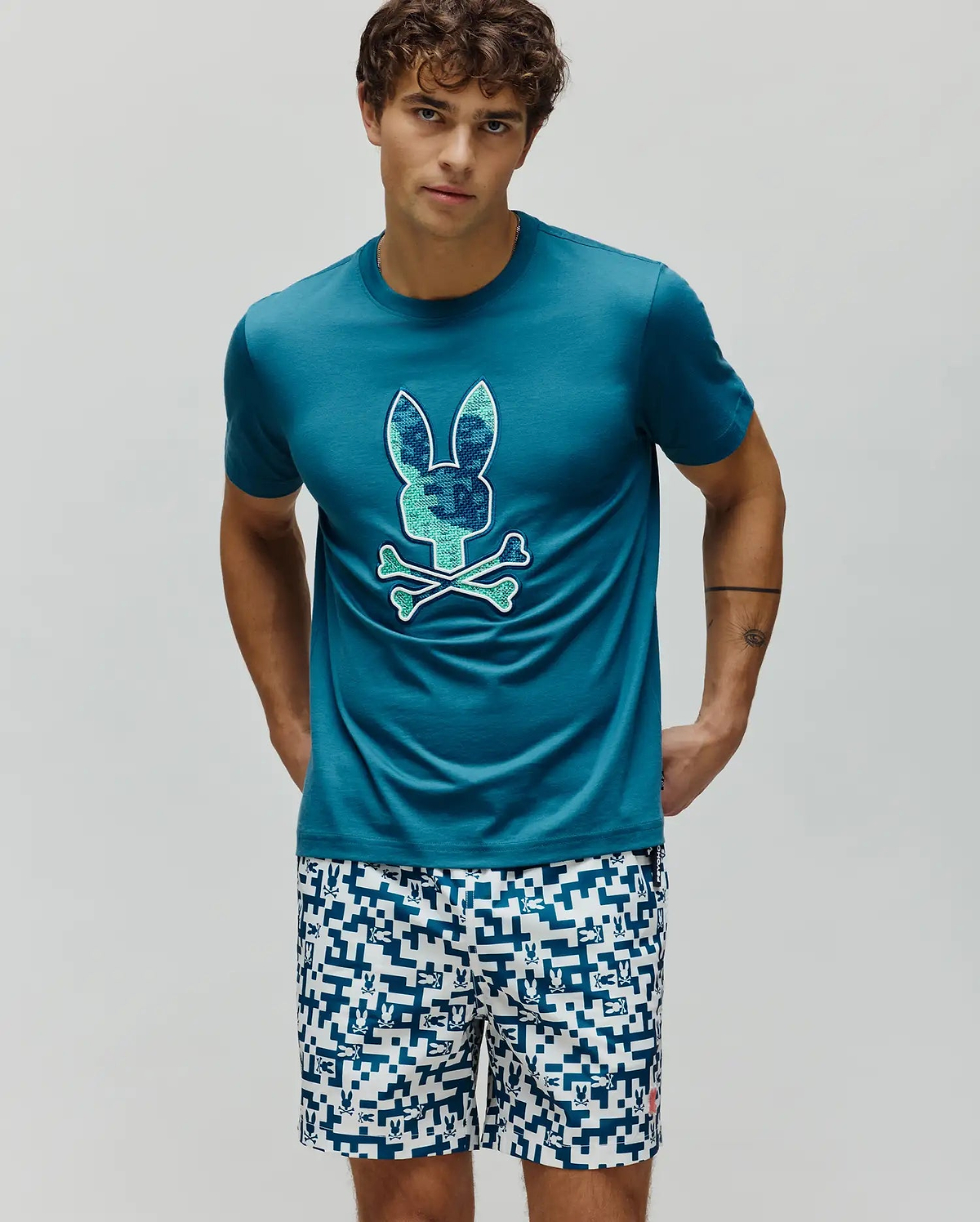 Mens Olivier Graphic Tee, a classic fit teal jersey t-shirt with a large pixelated blue and green Psycho Bunny skull and crossbones chest graphic. White shorts feature an all-over blue geometric bunny pattern. (2)