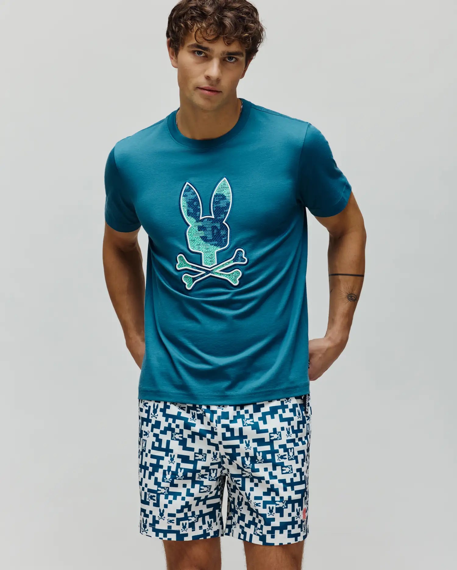 Mens Olivier Graphic Tee, a classic fit teal jersey t-shirt with a large pixelated blue and green Psycho Bunny skull and crossbones chest graphic. White shorts feature an all-over blue geometric bunny pattern. (2)