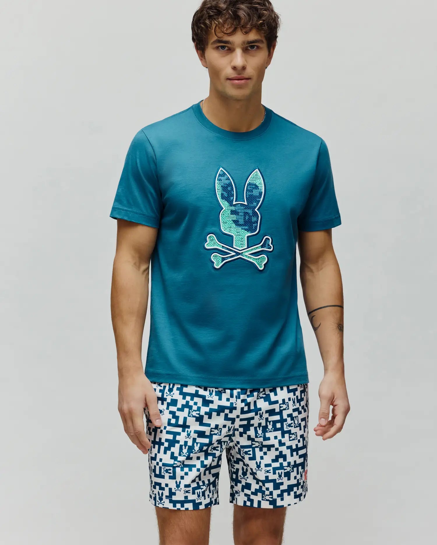 Mens Olivier Graphic Tee, a classic fit teal jersey t-shirt with a large, textured blue and green pixelated Psycho Bunny skull logo centered on the chest. (4)