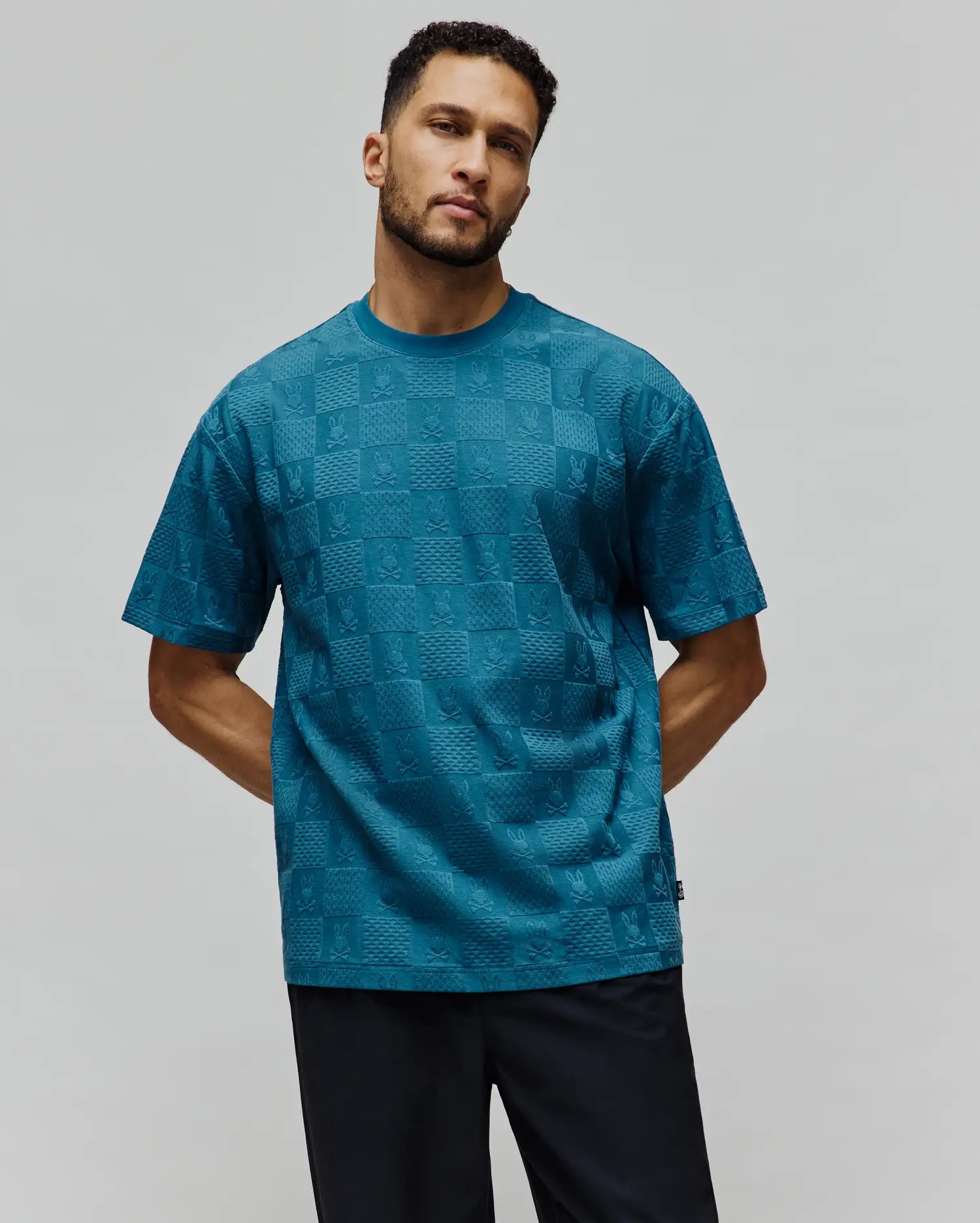 Mens Dustin Oversized Tee in teal, featuring an all-over embossed checkerboard pattern with alternating Psycho Bunny skull logos and textured squares, finished with a small brand tag. (4)