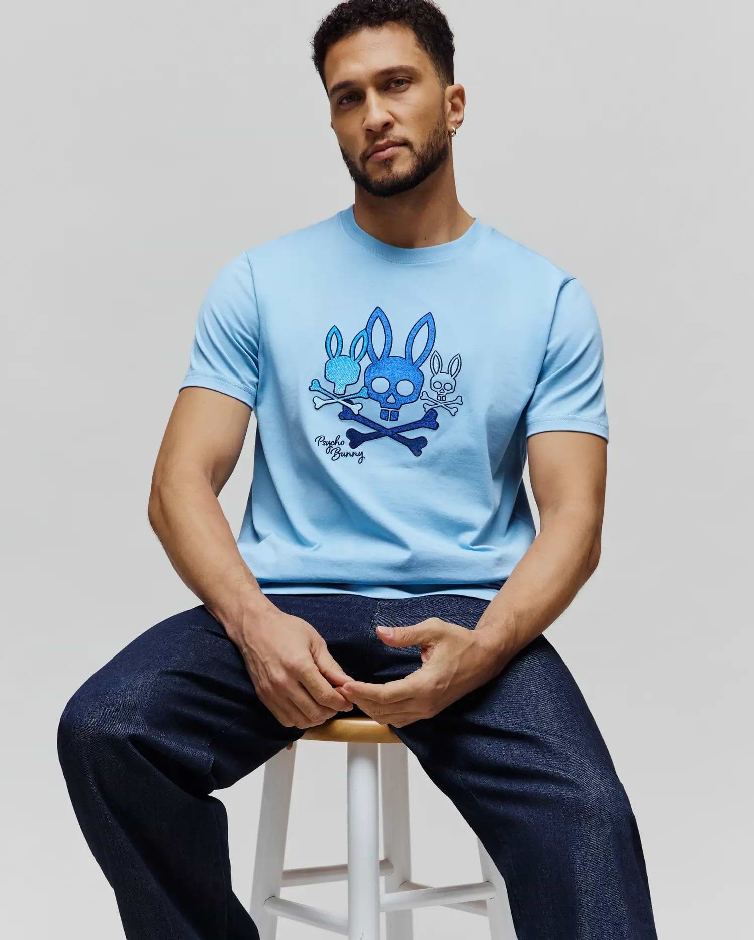 Mens Oscar Graphic Tee, a sky blue classic fit jersey t-shirt featuring a prominent chest graphic of three blue Psycho Bunny skull logos with crossbones and signature script. (3)