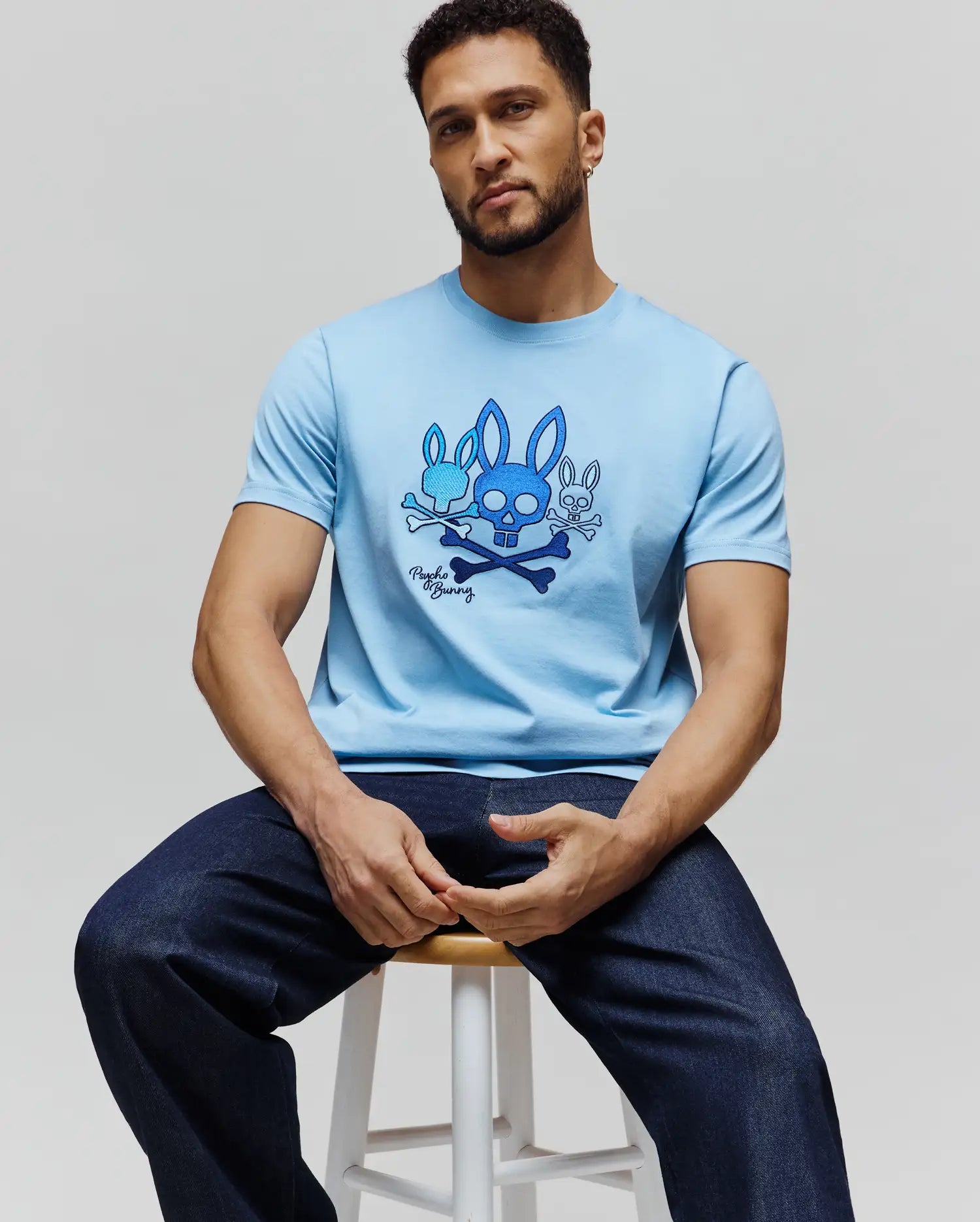Mens Oscar Graphic Tee, a sky blue classic fit jersey t-shirt featuring a prominent chest graphic of three blue Psycho Bunny skull logos with crossbones and signature script. (3)