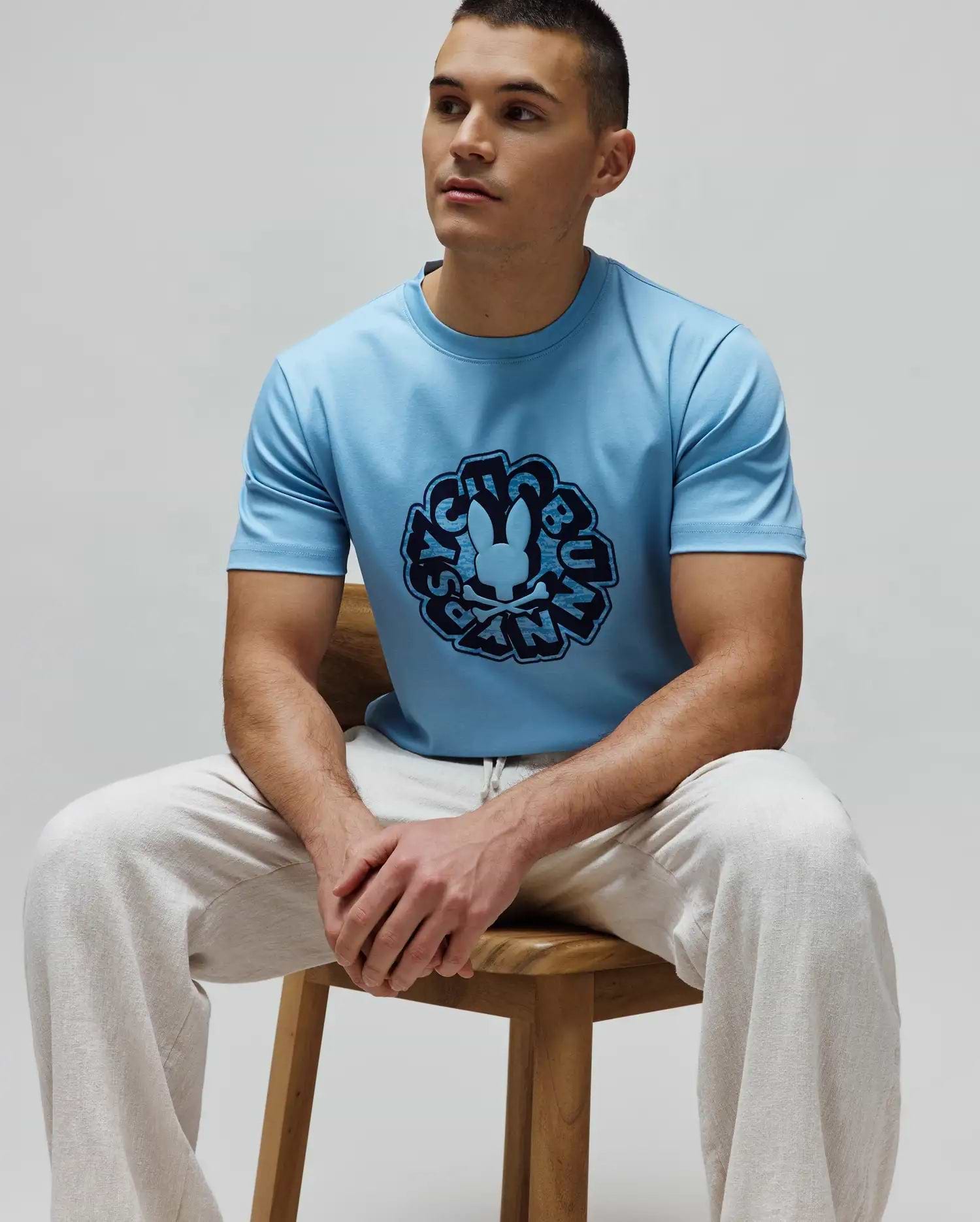 Mens Toby Graphic Tee in light blue, classic fit, smooth jersey knit, featuring a large dark blue circular Psycho Bunny skull logo and brand name graphic on the chest.