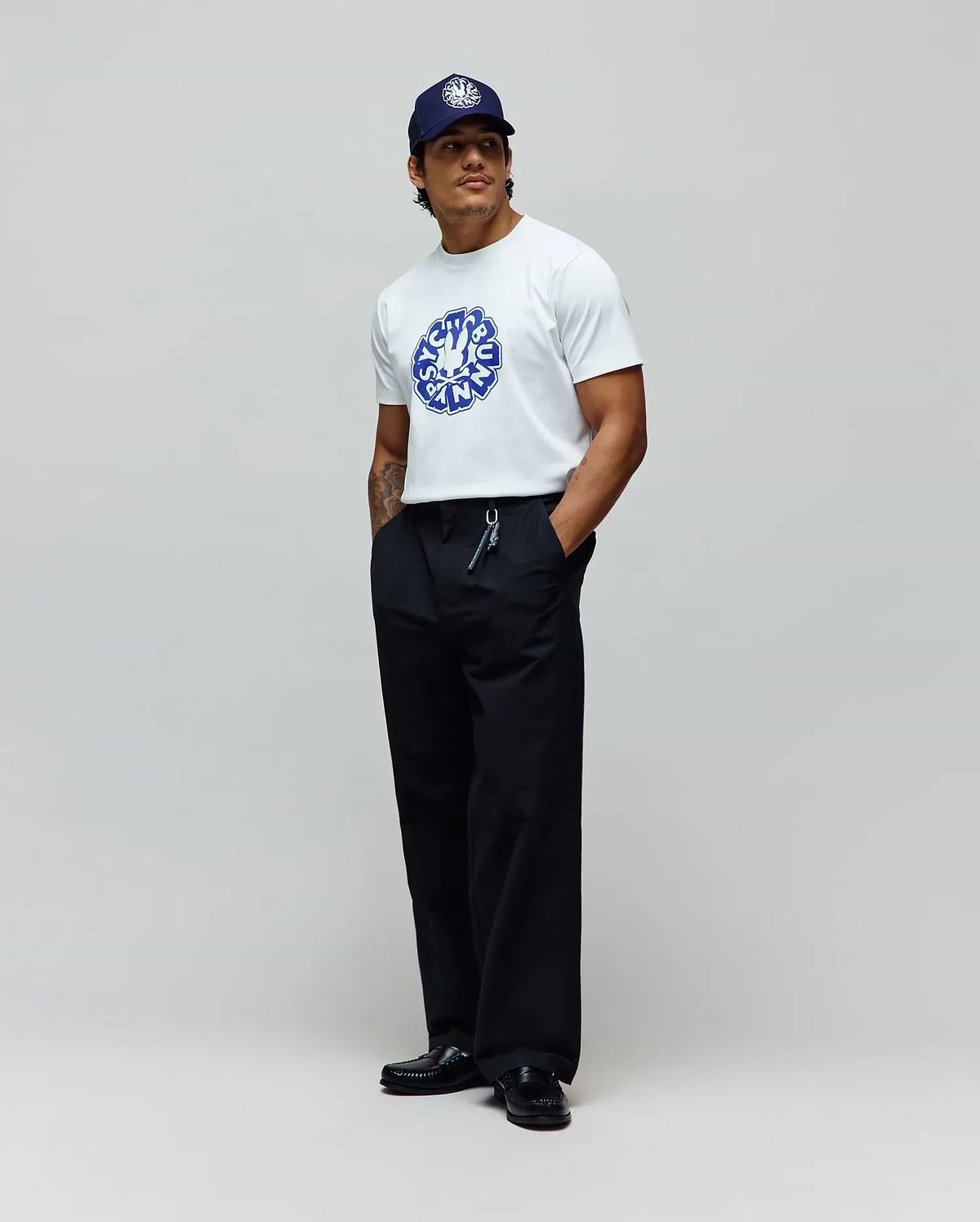 Mens Toby Graphic Tee, a white jersey t-shirt with a large blue Psycho Bunny circular graphic on the chest, paired with relaxed black pants and a navy cap featuring a white embroidered bunny skull. (3)