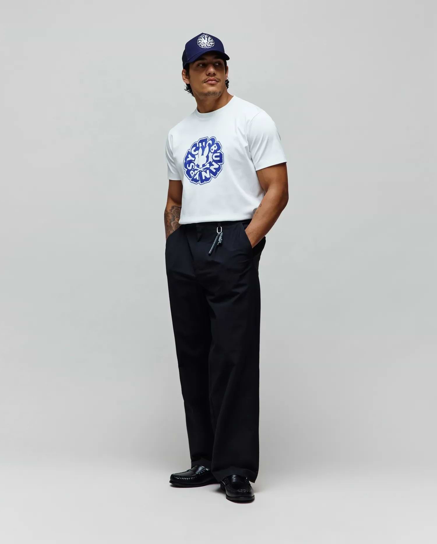 Mens Toby Graphic Tee, a white jersey t-shirt with a large blue Psycho Bunny circular graphic on the chest, paired with relaxed black pants and a navy cap featuring a white embroidered bunny skull. (3)
