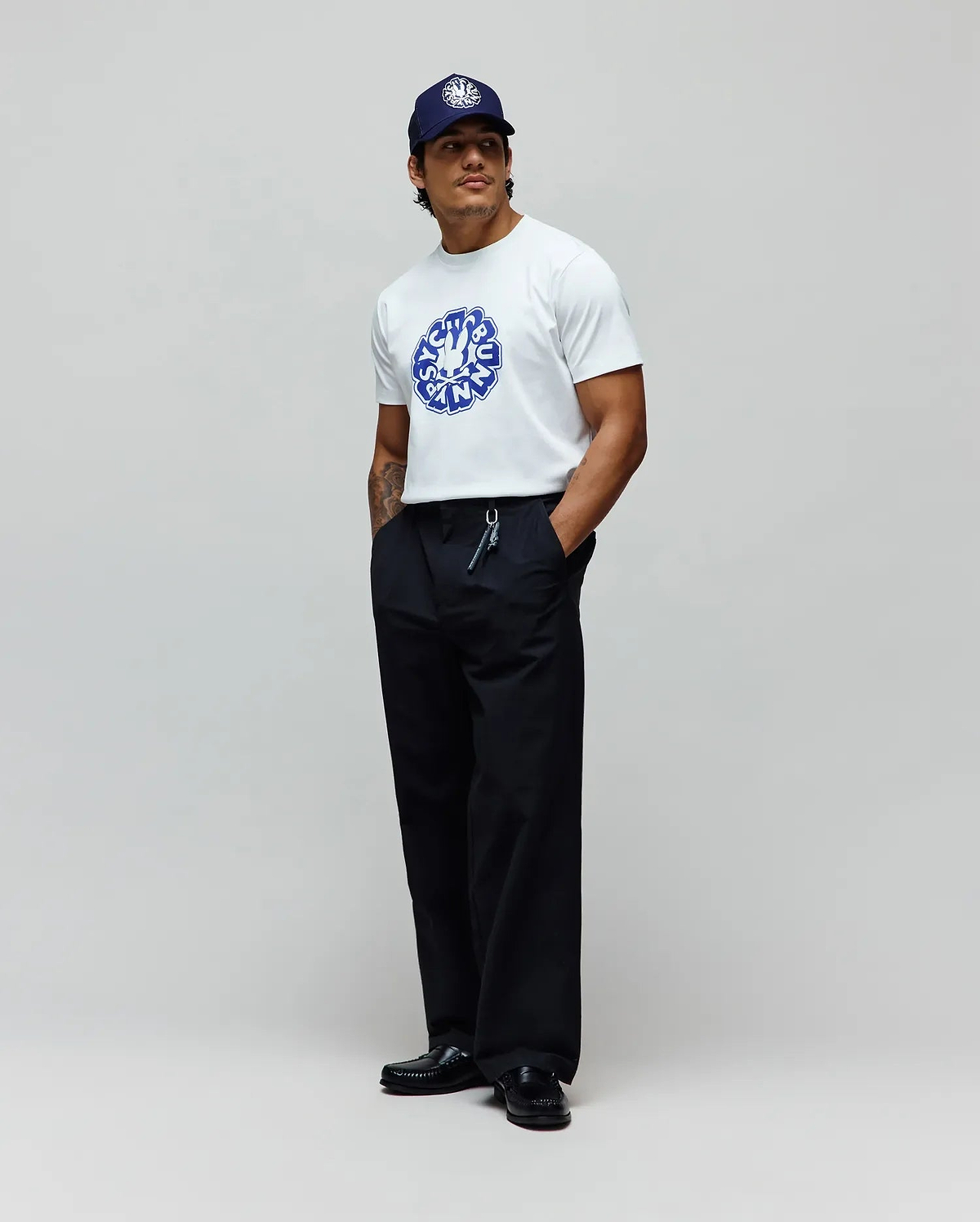 Mens Toby Graphic Tee, a white jersey t-shirt with a large blue Psycho Bunny circular graphic on the chest, paired with relaxed black pants and a navy cap featuring a white embroidered bunny skull. (3)