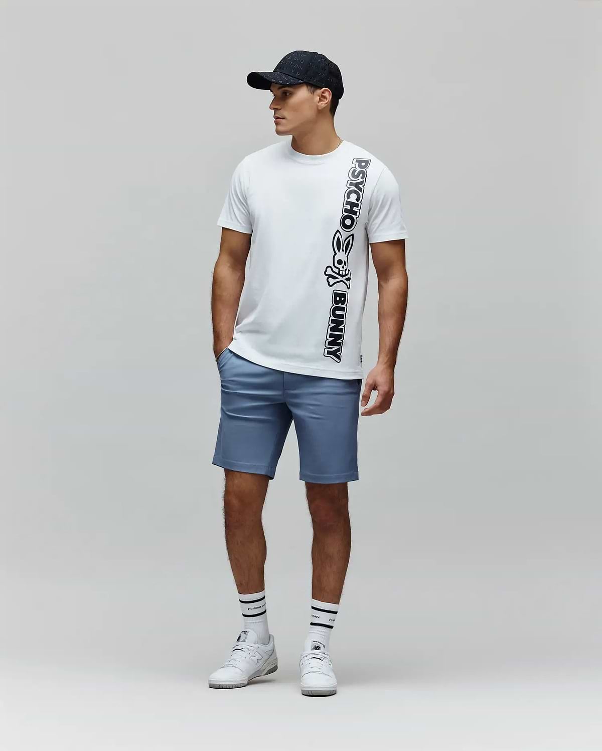 White Mens Toby Wordmark Tee, a classic fit jersey t-shirt, features a large vertical black Psycho Bunny skull logo and wordmark graphic on the front, paired with tailored blue shorts. (4)