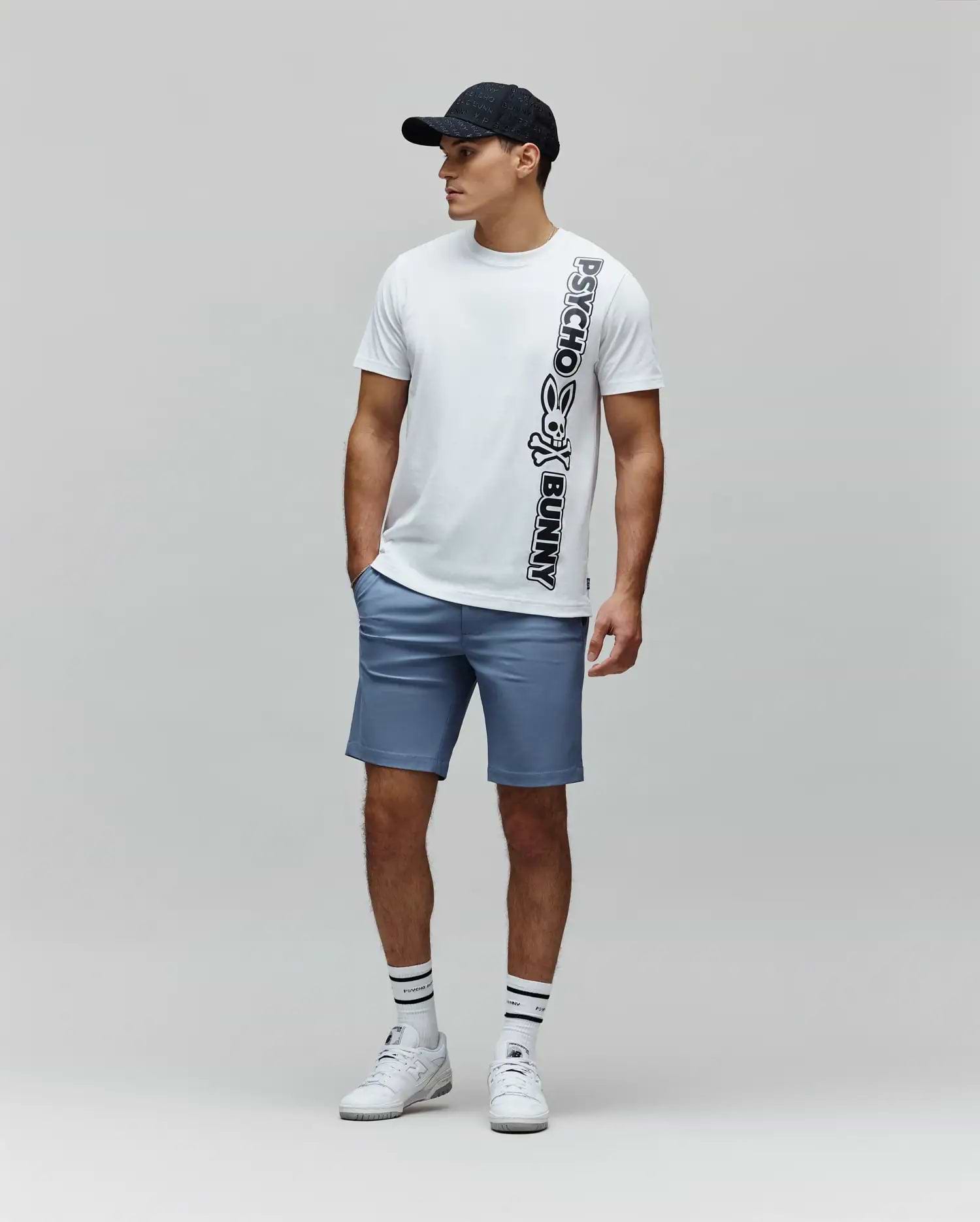 White Mens Toby Wordmark Tee, a classic fit jersey t-shirt, features a large vertical black Psycho Bunny skull logo and wordmark graphic on the front, paired with tailored blue shorts. (4)