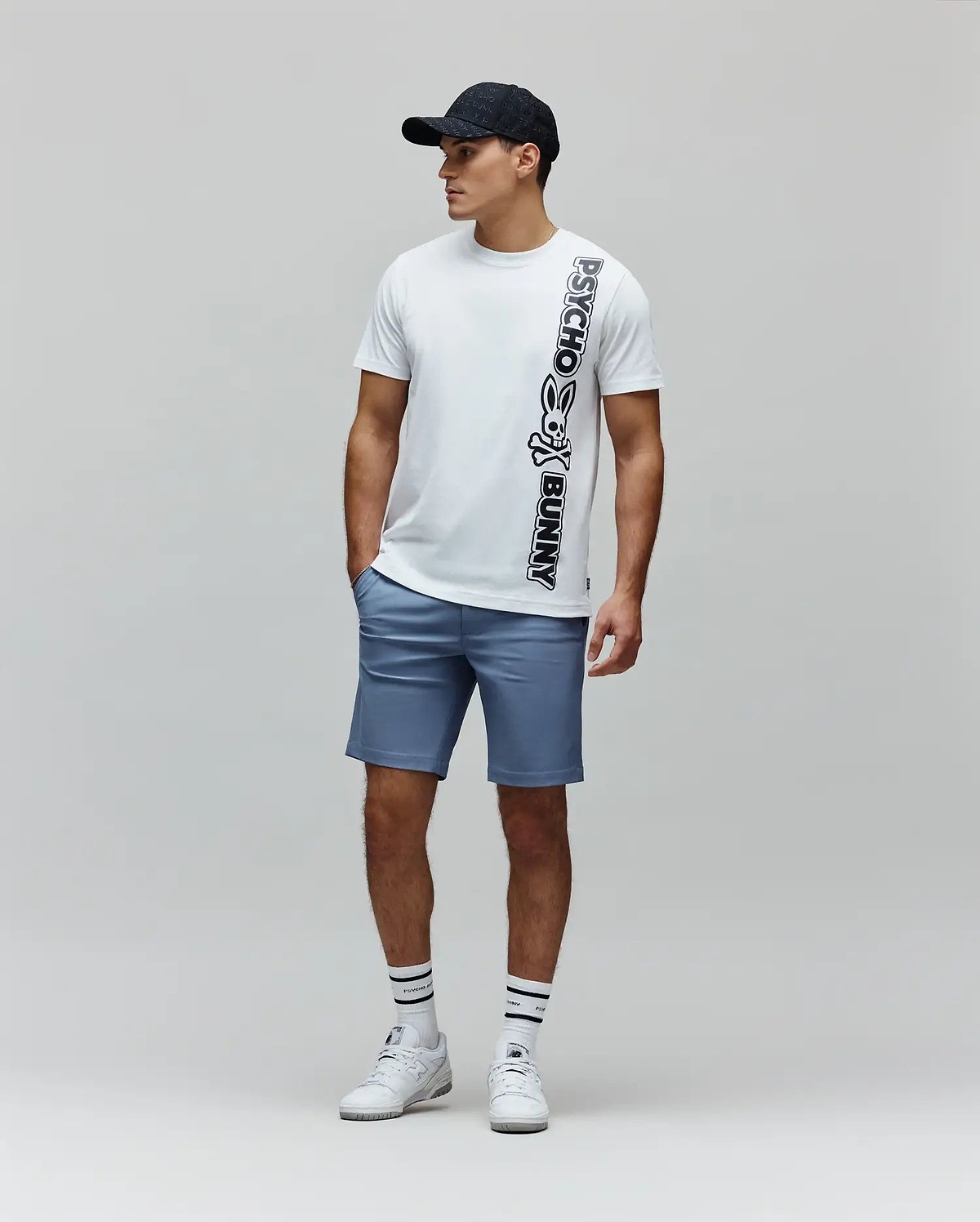 White Mens Toby Wordmark Tee, a classic fit jersey t-shirt, features a large vertical black Psycho Bunny skull logo and wordmark graphic on the front, paired with tailored blue shorts. (4)