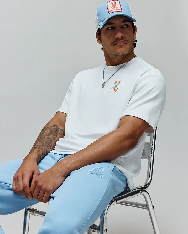 White Mens Ara Oversized Tee features a colorful deconstructed Psycho Bunny skull on the left chest, with light blue sweatpants with tonal logo and a light blue trucker cap with red brand patch.