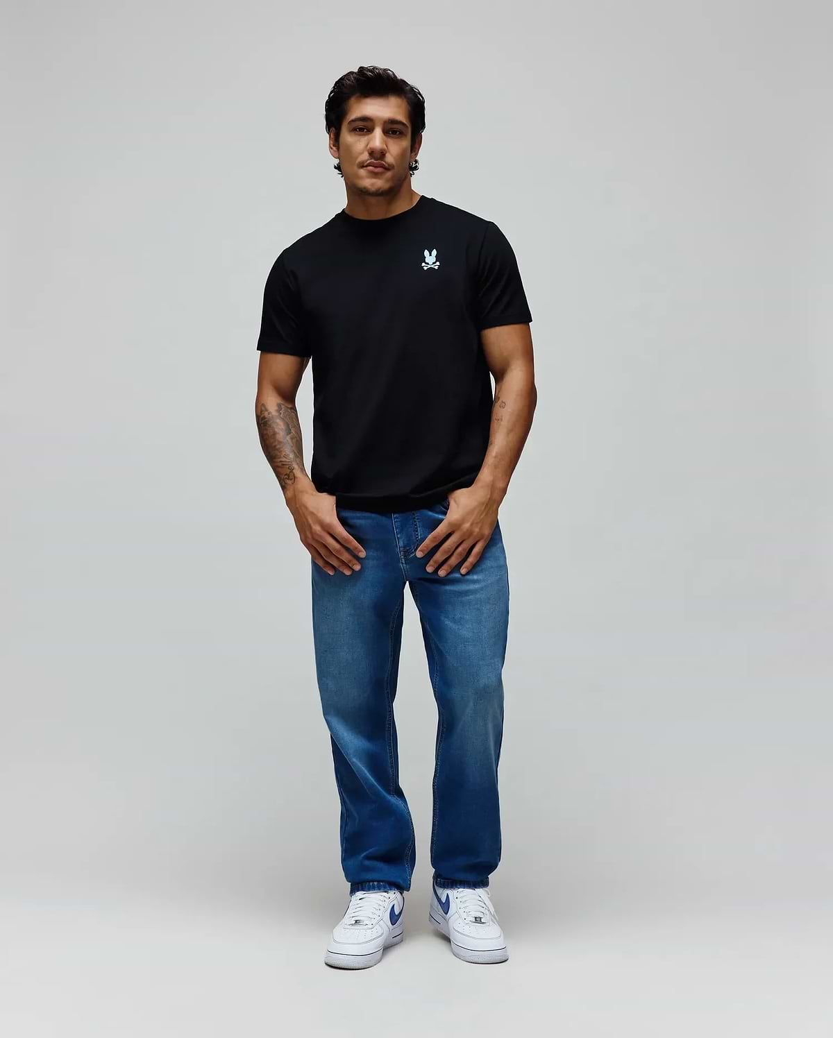 Black Mens Aiden Back Graphic Tee in classic fit jersey with a small light blue embroidered Psycho Bunny skull logo on the left chest, paired with relaxed fit medium wash blue denim jeans. (4)