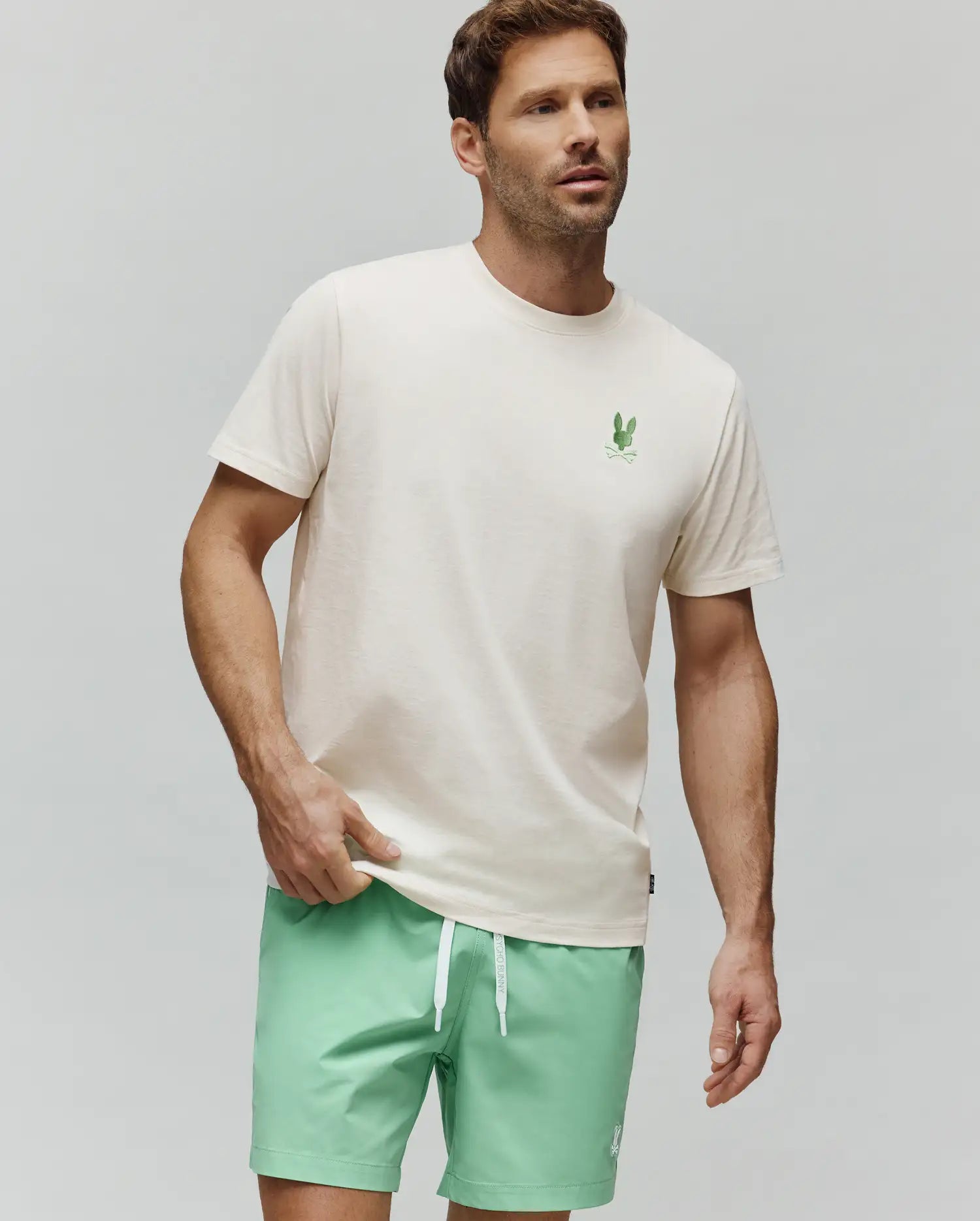 MENS AIDEN BACK GRAPHIC TEE in cream, crafted from soft jersey knit cotton with a classic fit. Features a small, vibrant green embroidered Psycho Bunny skull logo on the left chest and an implied back graphic. (3)