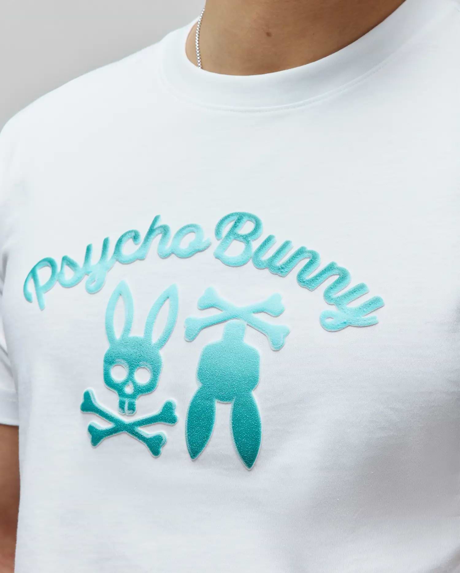 Mens Aram Graphic Tee, a white classic fit jersey t-shirt featuring a large central teal flocked Psycho Bunny script logo and deconstructed bunny skull graphic. (3)