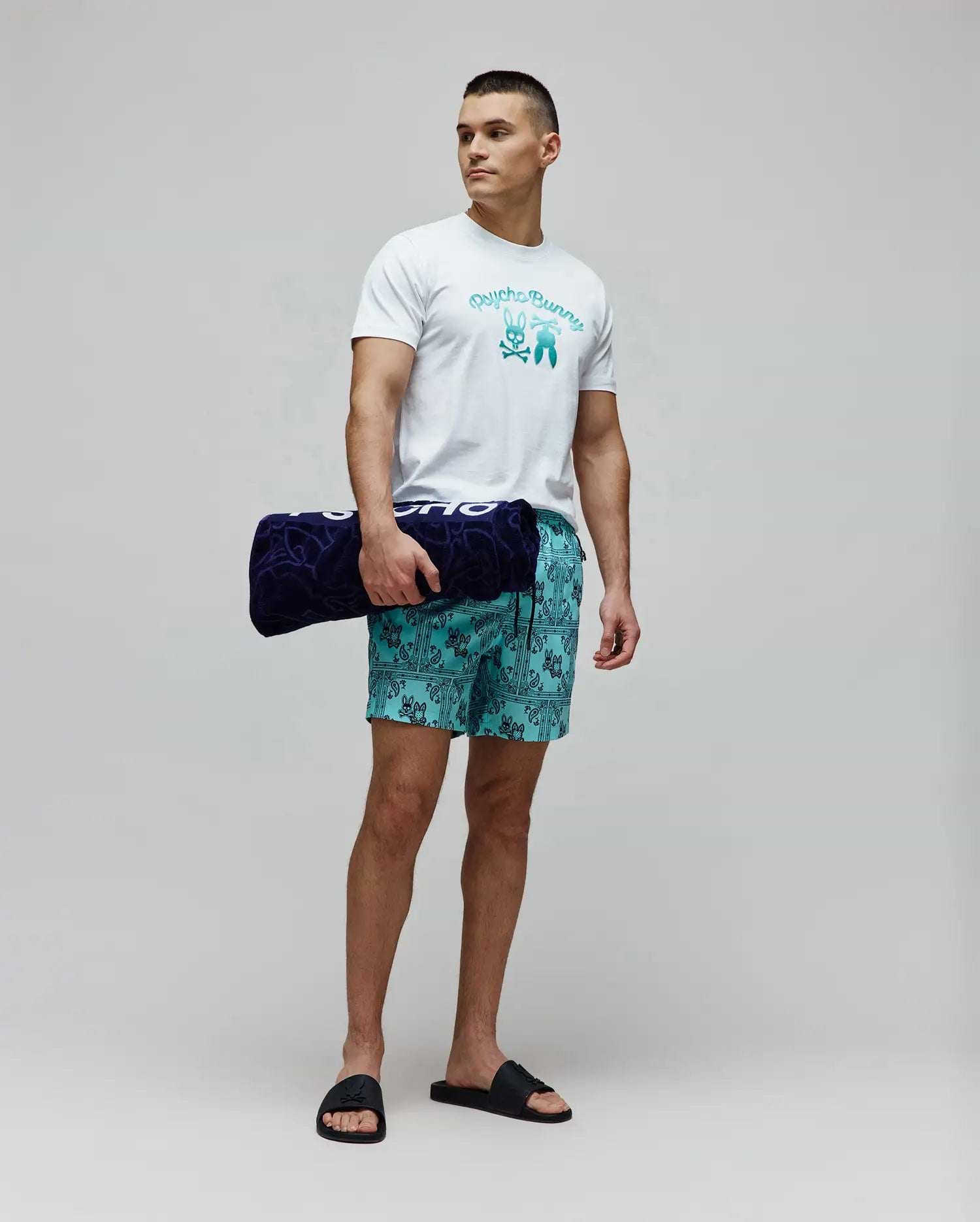Mens Aram Graphic Tee, white jersey with a large teal gradient Psycho Bunny skull logo, paired with teal bandana print swim shorts featuring small bunny skulls, and black logo slides. (4)