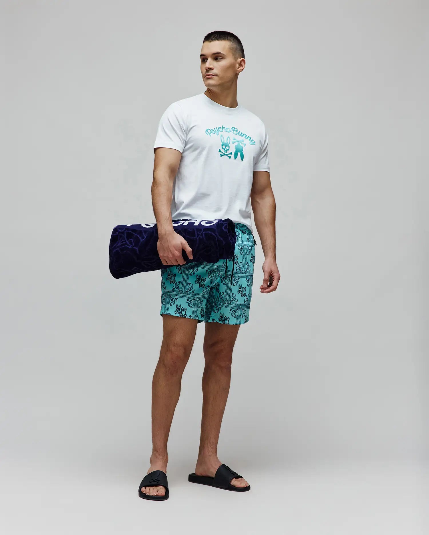 Mens Aram Graphic Tee, white jersey with a large teal gradient Psycho Bunny skull logo, paired with teal bandana print swim shorts featuring small bunny skulls, and black logo slides. (4)