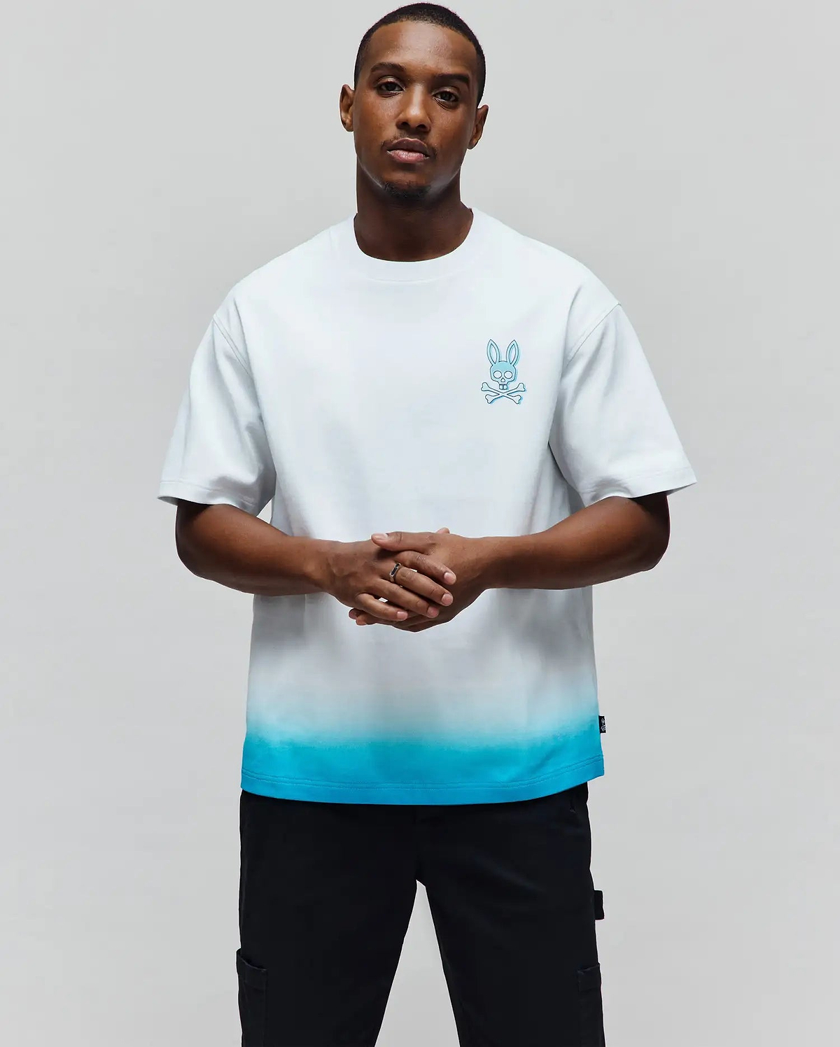 Mens Dax Oversized Tee, a white jersey t-shirt with an oversized fit, featuring a vibrant blue ombre fade at the hem and a small embroidered Psycho Bunny skull logo on the left chest. (1)