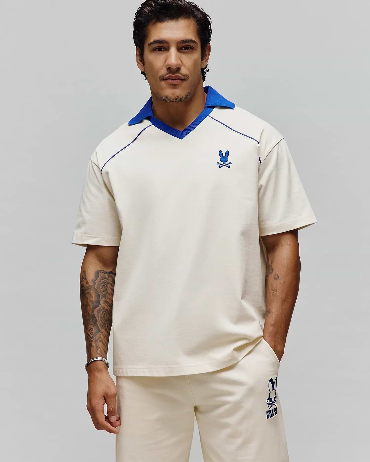Mens Toby Jersey, a relaxed fit cream jersey polo with a royal blue V-neck collar, blue piping, and an embroidered Psycho Bunny logo on the left chest. (1)